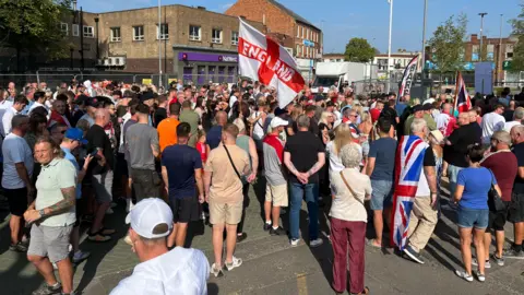 A crowd of people gathered on a street, with one St George's Cross flag being flown and one of the people wearing a Union Flag cape.