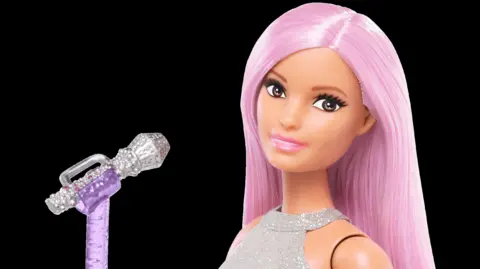 Mattel Inc Close-up of a fashion doll with long straight pink hair and a silver sleeveless top, posed beside a glittery purple microphone on a stand against a black background.