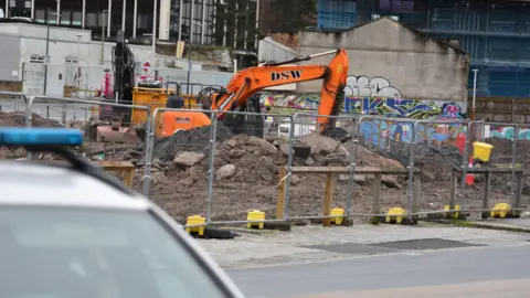 The image shows an orange digger at a construction site cordoned off with metal fencing. A police car is in the foreground. Mounds of earth and rubble are next to the digger.