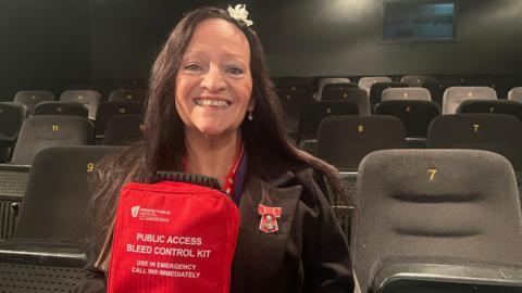 Lynne Baird holding a red bleed kit - it is labelled "public access bleed control kit. use in emergency."