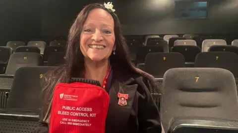 BBC Lynne Baird holding a red bleed kit - it is labelled "public access bleed control kit. use in emergency."