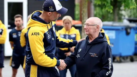Ed Sykes/Getty Images Former Leeds Rhinos' head coach Rohan Smith (left, wearing yellow and navy) shakes hands with Geoff Burrow (right, wearing a navy 'Team MND' hoody), the father of former Leeds Rhinos rugby league player and MND sufferer Rob Burrow, who died in summer 2024. Several other players are lined up in the background before they greet Geoff Burrow