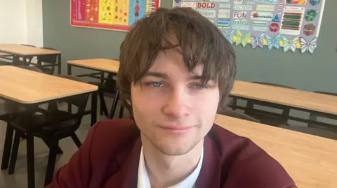Tiarnan has long brown hair that comes over one of his eyes. He is smiling slightly and has blue eyes. 