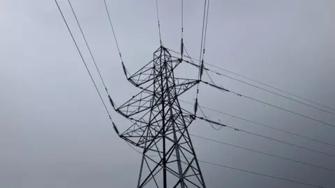 An electricity tower pictured silhouetted against a grey sky in Inverness. The structure is tall with metal lattice work and cables attached to it. 