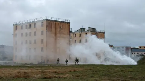 BBC Soldiers move towards a tall building as white smoke from a smoke grenade swirls around them