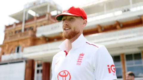 Ben Stokes is wearing a red cap and a white England kit