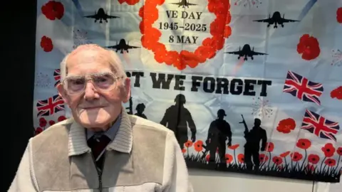 Kenneth Johnson is wearing glasses, looks at the camera, as he sits in front of large poster. The poster has a blue sky background with red poppies on it and silhoutees of soldiers, with British flags. It reads 'lest we forget' and 'VE DAY 1945-2025- 8 MAY