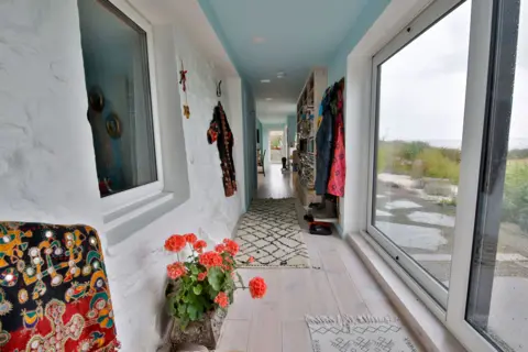 IWC Media A narrow, light-filled hallway features textured white walls, pale wood flooring, and a ceiling painted soft blue.
Large sliding glass doors line one side, while coats, bookshelves, and a patterned rug add colour and lived-in detail along the corridor.