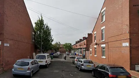 Google Cambridge Avenue, Whalley Range, Manchester, on a cloudy day showing a woman walking in the centre of the road with a walking stick and carrying a handbag.