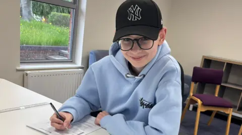 A boy with a pale blue hoodie, black cap and glasses sitting at a white desk, holding a pen over a sheet of paper with white walls and a window behind him