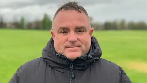 Sam Read/BBC Michael Mapp stands in a park with a grass field and slightly smiles at the camera. He has short dark hair and wears a black puffer coat. 