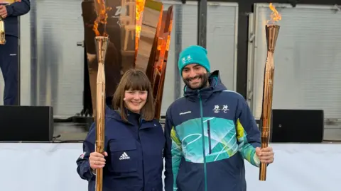 Millie Knight/King’s College London Two athletes, male and female, smile while holding golden torches topped with bright flames. Behind them is the Paralympic flame.