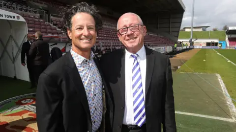Getty Images Brad Galinson and Paul Scally wearing suits and standing beside each other at Northampton Town Football Club.