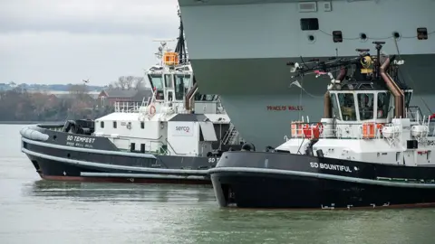 Serco Two tugs beside an aircraft carrier Prince of Wales