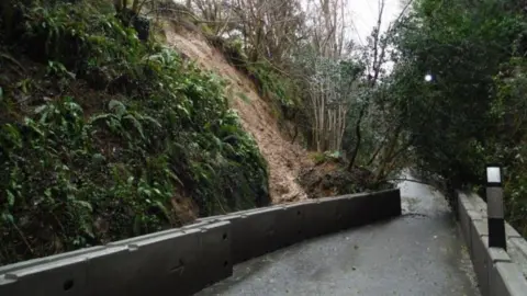 A road with a steep hill on one side, with a chunk of ground and vegetation having fallen towards the road and caused the concrete barriers lining the road to curve inwards over the tarmac, blocking the lane.