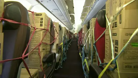 Boxes of disposable gloves strapped into seats of an aircraft. The overhead lockers are open.