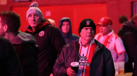 Getty Images A group of Southampton fans can be seen stood around in a stadium before a match. One of them in the foreground where's a club shirt and woolly hat and is holding a hot drink in a takeaway cup. 
