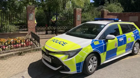 A police car with blue and yellow high-vis decals parked outside the metal gates of Queen's Park in Swindon. There are flowerbeds on either side of the gates and inside there is a paved path bordered with mature trees. Two police officers are standing monitoring the entrance.