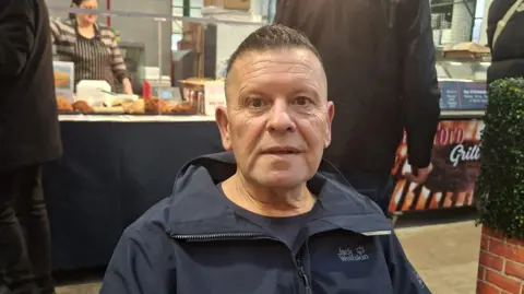 Thomas Kyle A man sitting outside in front of a food stall. He has short black hair and is wearing a navy jacket.