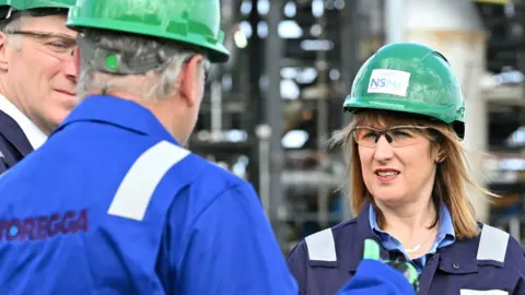 Getty Images Rachel Reeves, wearing an overall and helmet, talks to an official in a Storegga jacket in front of pipes.