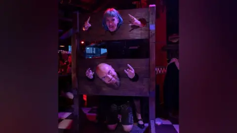 Lena Joyce A woman with blue hair is standing in medieval-style wooden stocks, with her head and both hands through circular holes. She has her tongue out and is doing "rock" signs with her hands. Below her is a man who is bald and has a long beard. He is on his knees with his head and both hands through the stocks' holes. He is smiling while holding his hands in "rock" signs