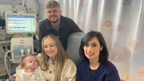 The Newcastle Upon Tyne Hospitals NHS Foundation Trust Poppy sits on her mother's knee while on a ward at the Freeman hospital. Poppy has a tube running from her nose. Her father stands behind her and Barbara Cardoso sits besides her.