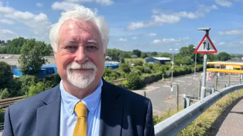 Steve Keable, Mid Devon District Council cabinet member for economic regeneration, in a light blue shirt, yellow tie and blue blazer, in the foreground with the railway line in the background.