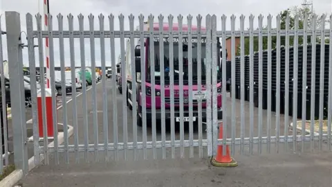 A purple waste lorry sits behind metal gates. An orange cone can be seen in front.