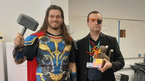 SBL Academy Two male teachers standing in a classroom with a whiteboard behind them. The man on the left is dressed as Thor and is holding a hammer, while the one on the right is Nick Fury wearing an eyepatch. He is holding a cat which has pipe cleaners coming out of its mouth.