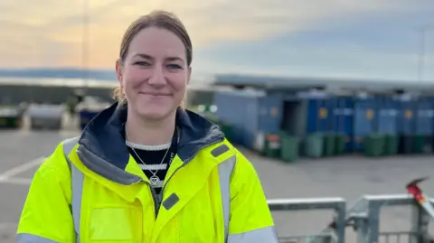 Sarah Robinson is standing in a yellow high viz coat and a striped black and white jumper is poking out underneath. Her light brown hair is tied back and she is smiling. She is standing on a balcony at the Longue Houge waste site, where recycling bins are in the blurred background of the image. The sky is a dusk light, with peach clouds and darkening blue.
