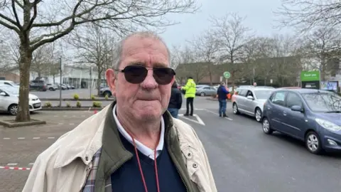 A white man with dark glasses wears a beige coat and black jumper. He looks at the camera. He is standing in Asda car park which has been turned into a bottled water station. Cars and people in high vis jackets are in the background.
