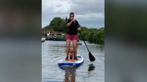 Help Our Canal A woman is standing on a Stund Up Paddling board in a canal with a dog by her feet.