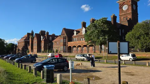 Paul Moseley/BBC A wide outdoor scene showing a large red-brick historic building with a clock tower. In front, a gravel car park contains several parked vehicles and a few people with a dog. Green grass and trees surround the area under bright, clear blue skies.