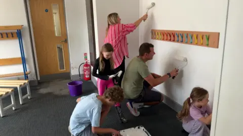 BBC Two adults and three children are all painting the same wall in a school corridor. Colourful coat hooks are in the middle of the wall and small wooden benches can be seen on the left. 