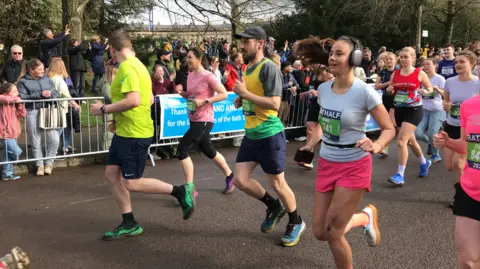 Runners wearing bibs reading Bath Half are mid-race, watched by lots of spectators behind short metal fencing. The runners are in bright athletic clothes.