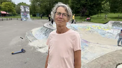 A woman wearing a pink t-shirt stares straight at the camera. She is wearing glasses and her hair is tied back. Behind her is a skatepark, including a steep skate ramp. It is covered in grafitti. A blue child's scooter is on the woman's left hand side behind her.