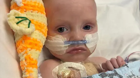 Macs Green is a small baby. He is lying in a hospital bed with a tube coming from his nose. He has an orange cuddly toy on his right