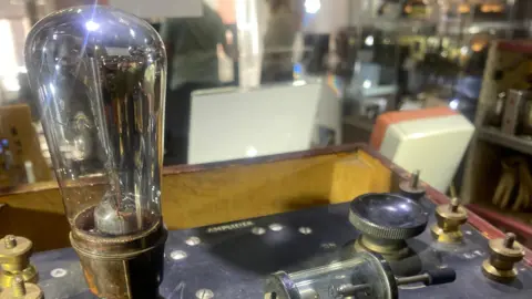 Daventry Museum Display case, with a glass valve on top of a Bakelite box on which are various metal fittings. The word AMPLIFIER can be seen on one edge of the box. There are other artefacts behind including boxes.