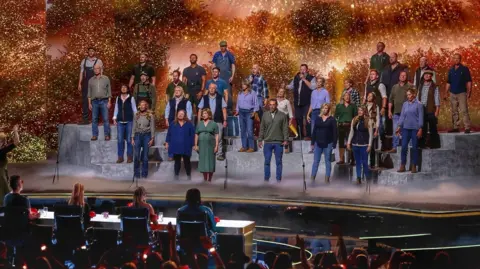 A large group of performers stands on tiered platforms on a brightly lit stage, appearing to sing together as a choir. A conductor stands at the front, guiding the performance. In front of the stage, a panel of judges sits at a table, watching. The background features warm, glowing lights and foliage-like scenery, while an audience in the foreground raises hands and watches the show.