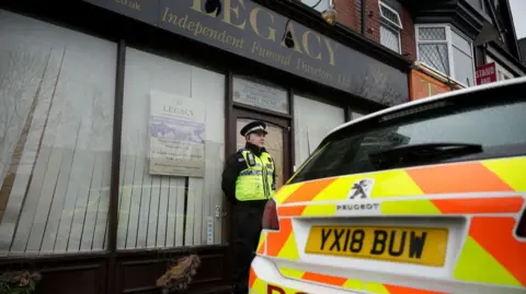 Getty Images A police officer standing outside Legacy Independent Funeral Directors in Hull. There is the rear of a police car in view and blinds on the windows of the premises