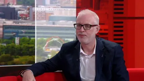 Paul is wearing a white shirt and navy blue suit jacket. He has short white hair and facial hair and is wearing black glasses with a red stripe.