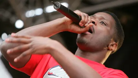 Lethal Bizzle holds a microphone to his mouth as he performs on stage at Love Music, Hate Racism in Trafalgar Square in 2006 - wearing a red t-shirt