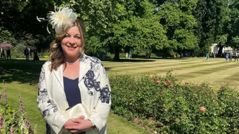 A lady is smiling at the camera. She is smartly dressed in a cream blazer with navy flowering detailing, and also has a matching cream fascinator on her head. She is stood in front of lush greenery of trees, hedges and a well-manicured lawn.