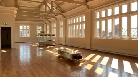 Milly Harrold/BBC Well lit large space with wooden floors and wide windows. An empty glass case is in the background, another one is in the foreground