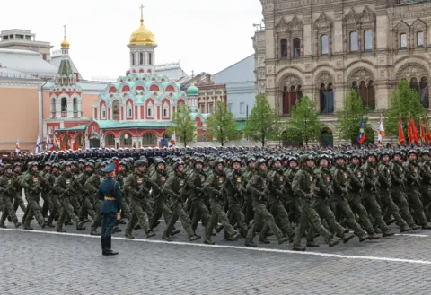 MAXIM SHIPENKOV/EPA/Shutterstock Russian servicemen march in rows wearing dark green fatigues, helmets with goggles placed on top, and holding guns. Kazan Cathedral, which is painted teal, red and white and features a gold dome, is behind them.