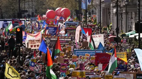 A crowd of people walk London, their heads are seen amongst dozens of colourful signs, flags and plaquards.