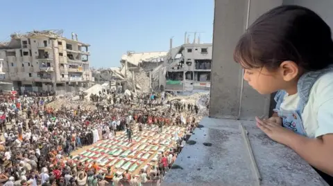 BBC A girl watches the mass funeral of 112 members of the Hassayna and Abu Sharia clans who the Civil Defence says were killed in an Israeli strike in November 2023, in Gaza City, northern Gaza (4 August 2026)