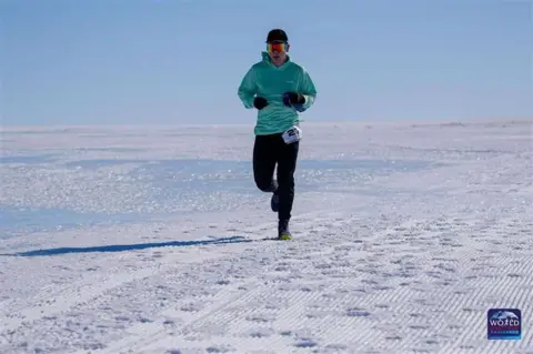 Mr Holborn, wearing an orange sun visor, teal hoodie and black trousers, running along the ice of Antarctica