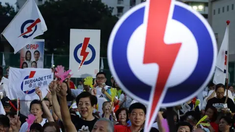 Getty Images People's Action Party supporters in Singapore cheer at an election rally, with many holding up posters and flags with the party symbol of a red lightning bolt in a blue and white circle. In the foreground is a large lit sign of the PAP symbol