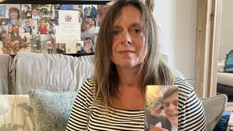 Bridget Lucas sitting on a sofa in a black and white striped top holding a photograph of her son Lawrence. A notice board behind her has more family photos.